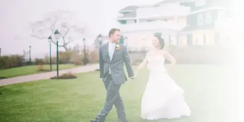 A bride and groom walking hand-in-hand on a grassy lawn in front of a large house.
