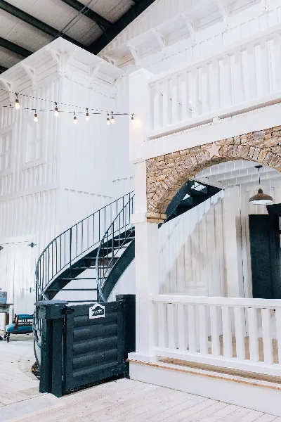 Interior view of a rustic event venue featuring a black spiral staircase, white wooden walls, and a stone archway.