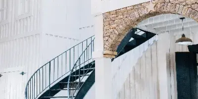 Interior view of a rustic event venue featuring a black spiral staircase, white wooden walls, and a stone archway.