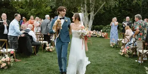 A bride and groom walking down the aisle during an outdoor wedding ceremony with guests seated on either side.