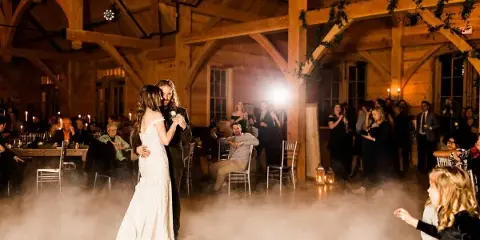 A wedding reception in a wooden barn with set dining tables and a couple dancing.