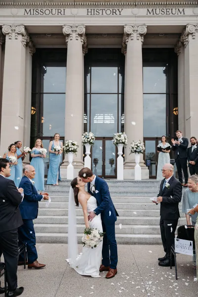 A bride and groom kissing on the steps of the Missouri History Museum while guests throw petals.