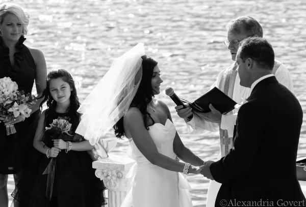 A black and white photo of a wedding officiant performing a ceremony for a bride and groom outdoors near a body of water.