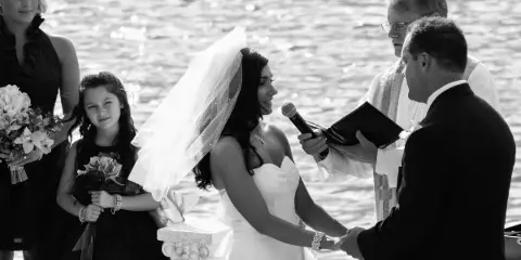A black and white photo of a wedding officiant performing a ceremony for a bride and groom outdoors near a body of water.