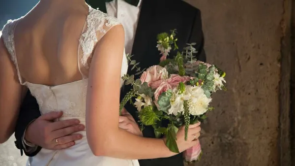 A bride in a white lace dress and a groom in a dark suit standing together, with the bride holding a bouquet of pink and white flowers.