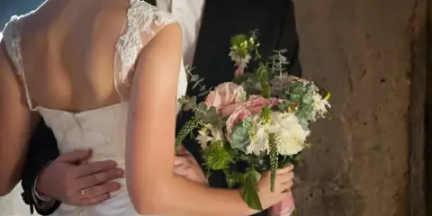 A bride in a white lace dress and a groom in a dark suit standing together, with the bride holding a bouquet of pink and white flowers.