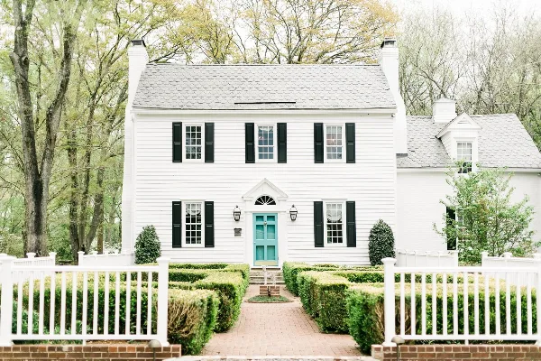 Exterior view of a white historic two-story manor house with black shutters, a teal front door, and a white picket fence.