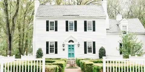Exterior view of a white historic two-story manor house with black shutters, a teal front door, and a white picket fence.
