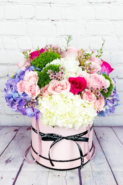 A vibrant floral arrangement featuring pink roses, white hydrangeas, and purple orchids arranged in a pink cylindrical box with a black ribbon.