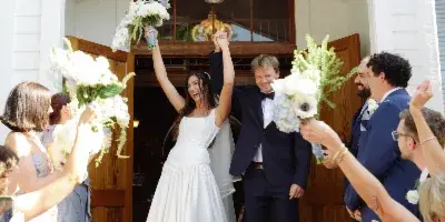 A bride and groom exit a white chapel with the number 1897 above the door, raising their bouquets and hands in celebration while surrounded by cheering guests.