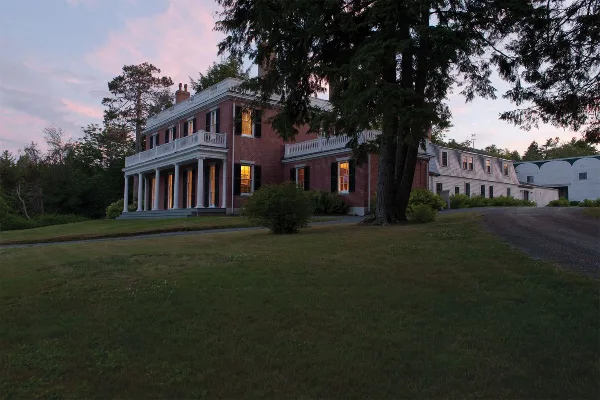 Historic red brick house with white columns and balconies on a grassy lawn at dusk, with a large tree in the foreground.