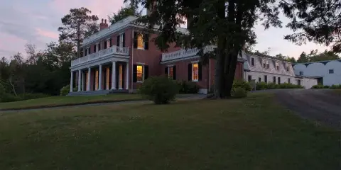 Historic red brick house with white columns and balconies on a grassy lawn at dusk, with a large tree in the foreground.