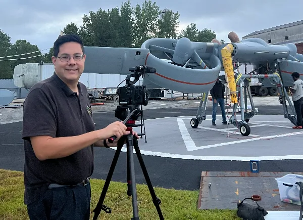 Justin Chan, owner of Valley Creek Productions, standing with a professional video camera on a tripod in front of an aircraft.
