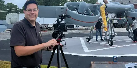 Justin Chan, owner of Valley Creek Productions, standing with a professional video camera on a tripod in front of an aircraft.