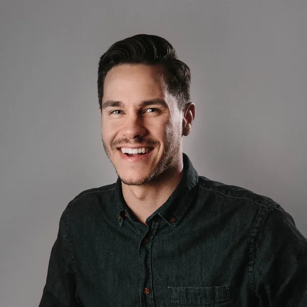 Portrait of a smiling man with dark hair and a beard wearing a dark button-down shirt against a gray background.