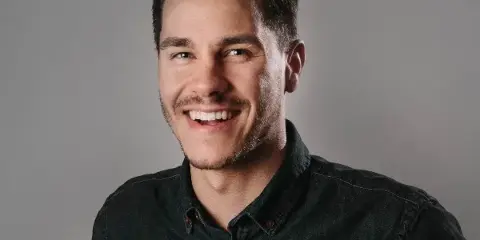 Portrait of a smiling man with dark hair and a beard wearing a dark button-down shirt against a gray background.
