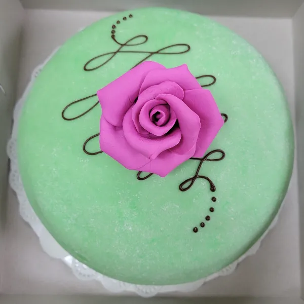 A round cake with green frosting, decorated with a pink fondant rose and chocolate drizzle.