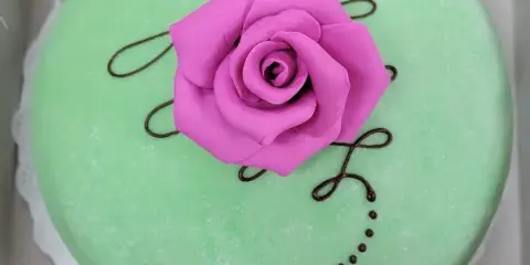 A round cake with green frosting, decorated with a pink fondant rose and chocolate drizzle.