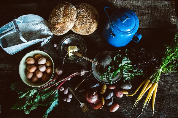Overhead view of rustic bread loaves, butter, eggs, and fresh vegetables arranged on a dark wooden table.