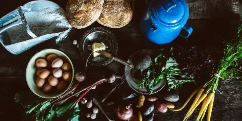 Overhead view of rustic bread loaves, butter, eggs, and fresh vegetables arranged on a dark wooden table.