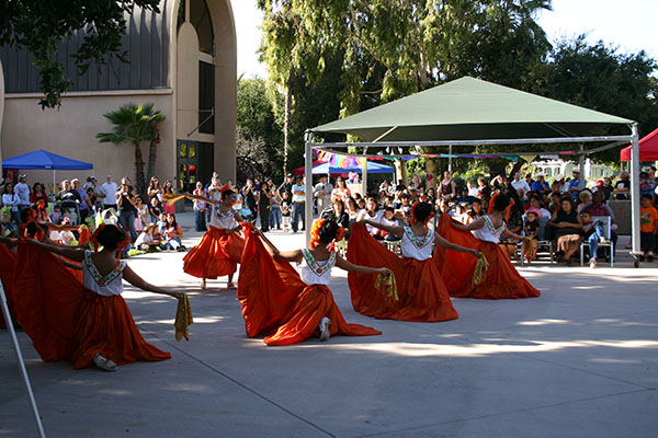 A group of dancers in red skirts performing in an outdoor courtyard in front of a seated audience.