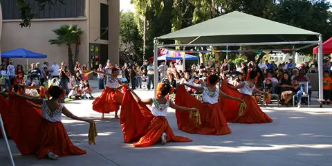 A group of dancers in red skirts performing in an outdoor courtyard in front of a seated audience.