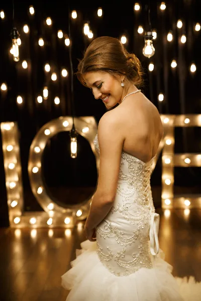 A bride in a white gown stands in front of a large illuminated marquee letter 'L' and hanging light bulbs.