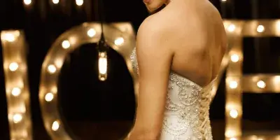 A bride in a white gown stands in front of a large illuminated marquee letter 'L' and hanging light bulbs.