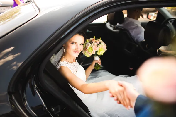 A bride in a white wedding dress holding a floral bouquet sits in the back seat of a black luxury car, reaching out to be helped by a person outside the vehicle.