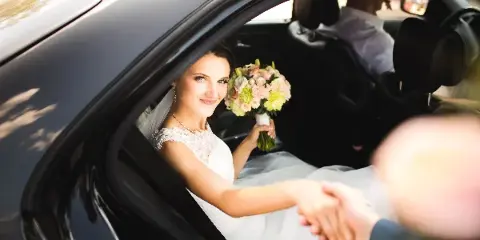 A bride in a white wedding dress holding a floral bouquet sits in the back seat of a black luxury car, reaching out to be helped by a person outside the vehicle.