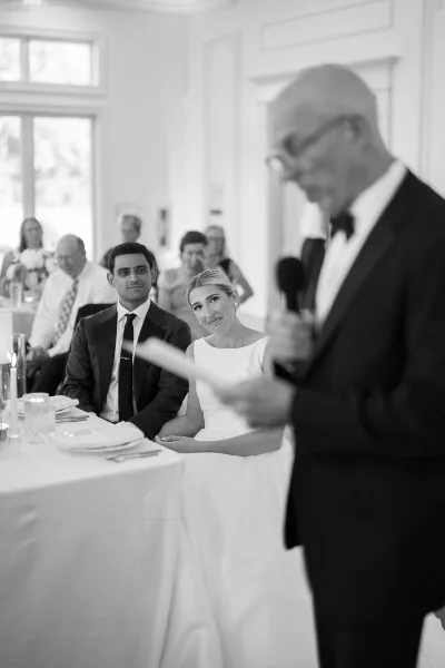 Black and white photo of a bride and groom seated at a table during a wedding reception while a man speaks into a microphone.