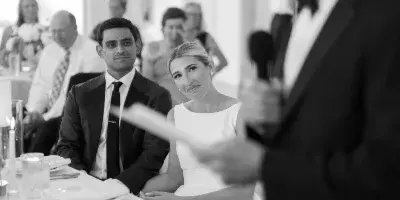 Black and white photo of a bride and groom seated at a table during a wedding reception while a man speaks into a microphone.