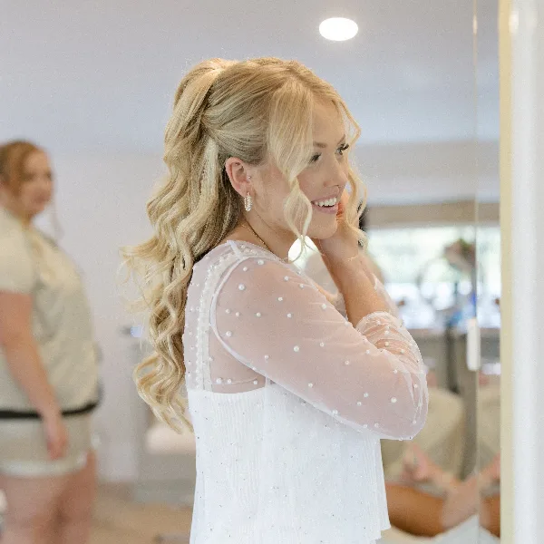 A smiling blonde bride with a wavy half-up hairstyle and makeup, wearing a white dress in a studio.