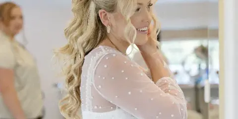A smiling blonde bride with a wavy half-up hairstyle and makeup, wearing a white dress in a studio.