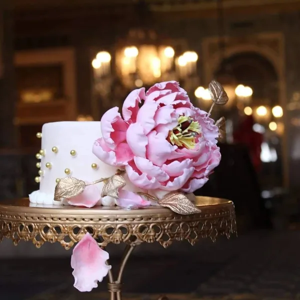 A white cake decorated with a large pink sugar peony, gold leaves, and gold bead accents, displayed on an ornate gold cake stand.
