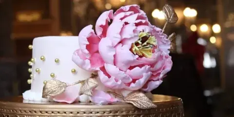 A white cake decorated with a large pink sugar peony, gold leaves, and gold bead accents, displayed on an ornate gold cake stand.