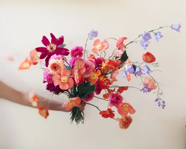 A hand holding a colorful, loose bouquet of garden flowers including ranunculus and cosmos against a neutral background.