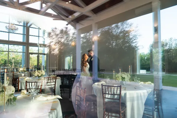 A bride and groom standing in a glass-walled wedding reception hall with set tables and outdoor views.