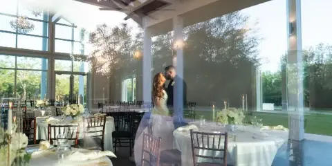 A bride and groom standing in a glass-walled wedding reception hall with set tables and outdoor views.