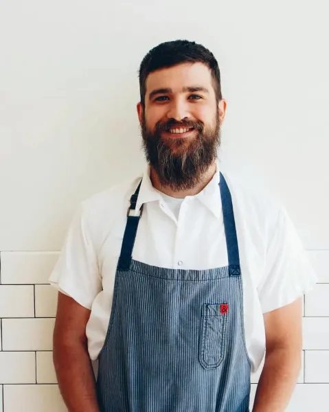 Portrait of a bearded chef wearing a white shirt and blue striped apron standing in a kitchen.