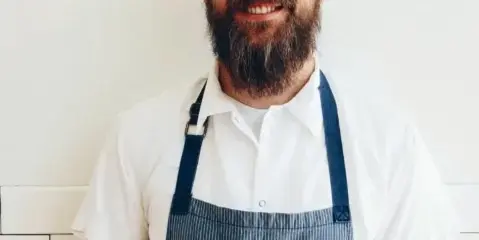 Portrait of a bearded chef wearing a white shirt and blue striped apron standing in a kitchen.