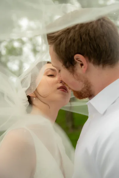 A close-up, artistic wedding photograph of a bride and groom facing each other, with the bride's veil draped over their heads.