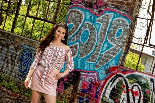 A professional portrait of a young woman with long brown hair wearing a striped top and pink skirt, posing in front of a brick wall with colorful graffiti.