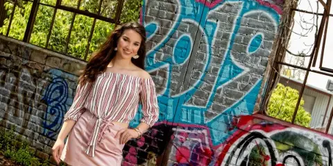 A professional portrait of a young woman with long brown hair wearing a striped top and pink skirt, posing in front of a brick wall with colorful graffiti.