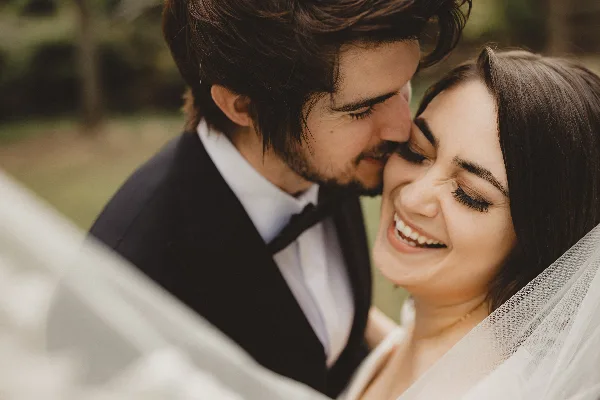 A candid, close-up photograph of a smiling bride and groom sharing an intimate moment outdoors.
