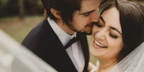A candid, close-up photograph of a smiling bride and groom sharing an intimate moment outdoors.