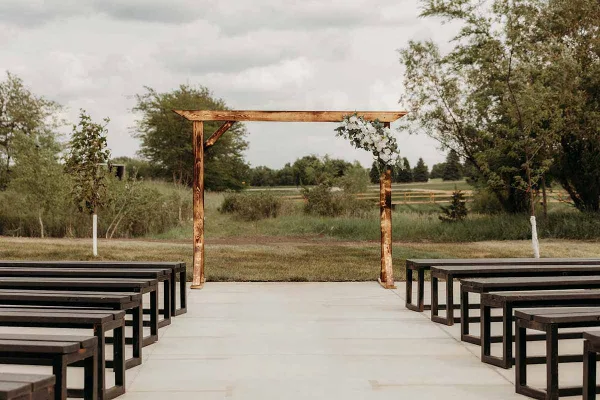 Outdoor wedding ceremony area featuring a wooden arch with white flowers and rows of dark wooden benches.