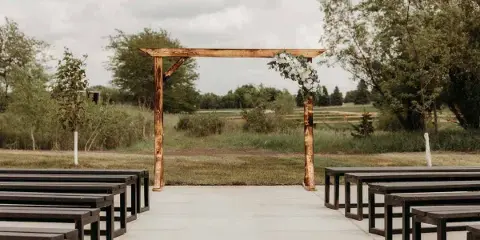Outdoor wedding ceremony area featuring a wooden arch with white flowers and rows of dark wooden benches.