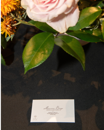 Close-up of a fresh floral arrangement with a pink rose and green leaves, with a Juliette Marie Floral Design business card in the foreground.