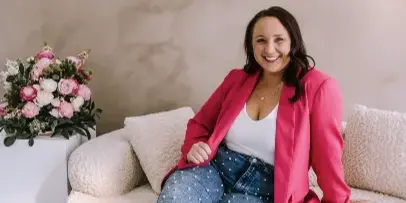 Smiling woman in a pink blazer seated on a cream sofa beside pink and white floral arrangements.
